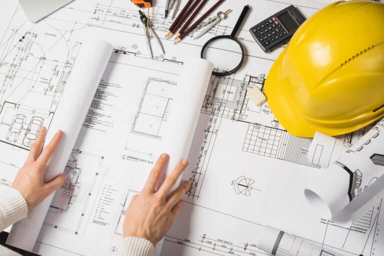 Hands holding rolled-up blueprints on a construction planning table with a yellow hard hat, magnifying glass, and drafting tools.